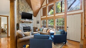 Amplia sala de estar con chimenea de piedra, vigas de madera y ventanas grandes que ofrecen vistas al exterior.