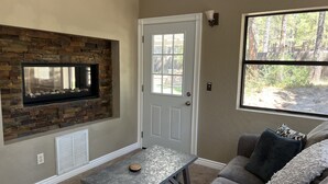 Ein Wohnzimmer mit Kamin aus Stein, einer weißen Tür, einem Fenster mit Blick auf Bäume und einem grauen Sofa mit Kissen.