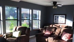 Ein Wohnzimmer mit zwei braunen Ledersofas, einem Deckenventilator und großen Fenstern mit Blick auf Grünflächen.