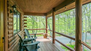 A wooden cabin with a porch, two Adirondack chairs, and a view of a forested area.