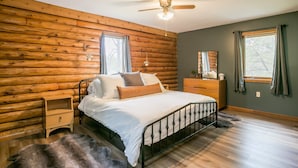 A bedroom with a wooden log wall, a bed with white bedding, a nightstand, a dresser, and a ceiling fan.
