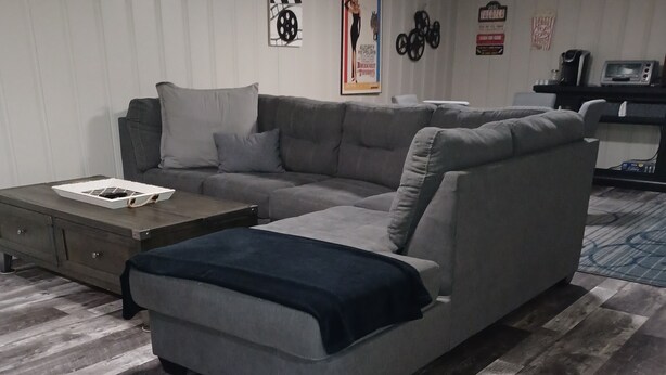 A living room with a sectional sofa, a coffee table, and a rug. There are wall decorations and a kitchen area in the background. - Spacious and Serene Basement (McMinnville)