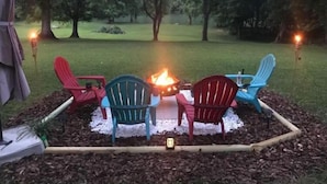 A fire pit with four Adirondack chairs around it, set in a garden with mulch and trees in the background. - Spacious and Serene Basement (McMinnville)