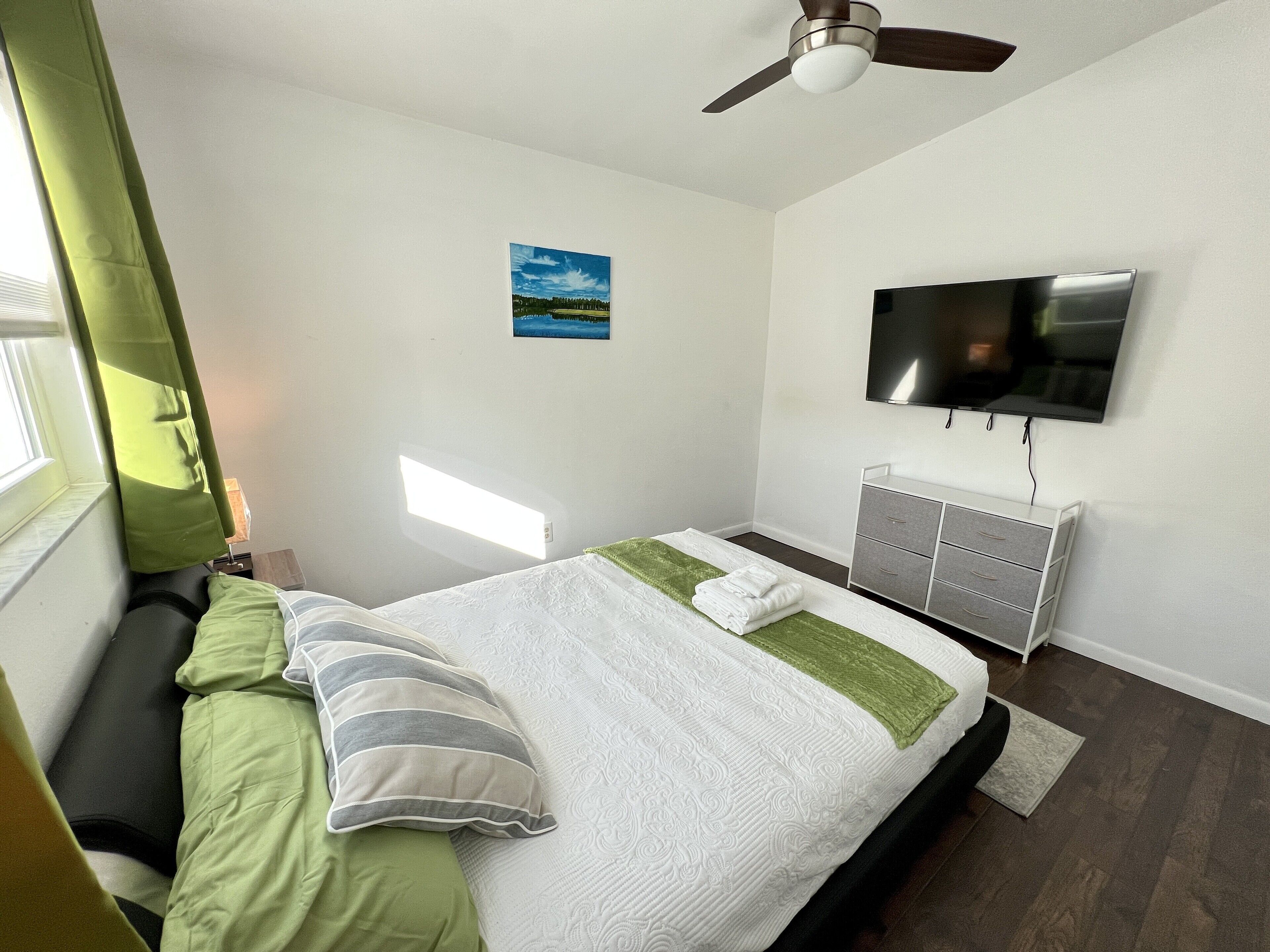 A bedroom with a bed, a TV, a ceiling fan, a window, and a painting on the wall.