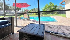 A covered patio area with a pool, wicker furniture, and a red umbrella.