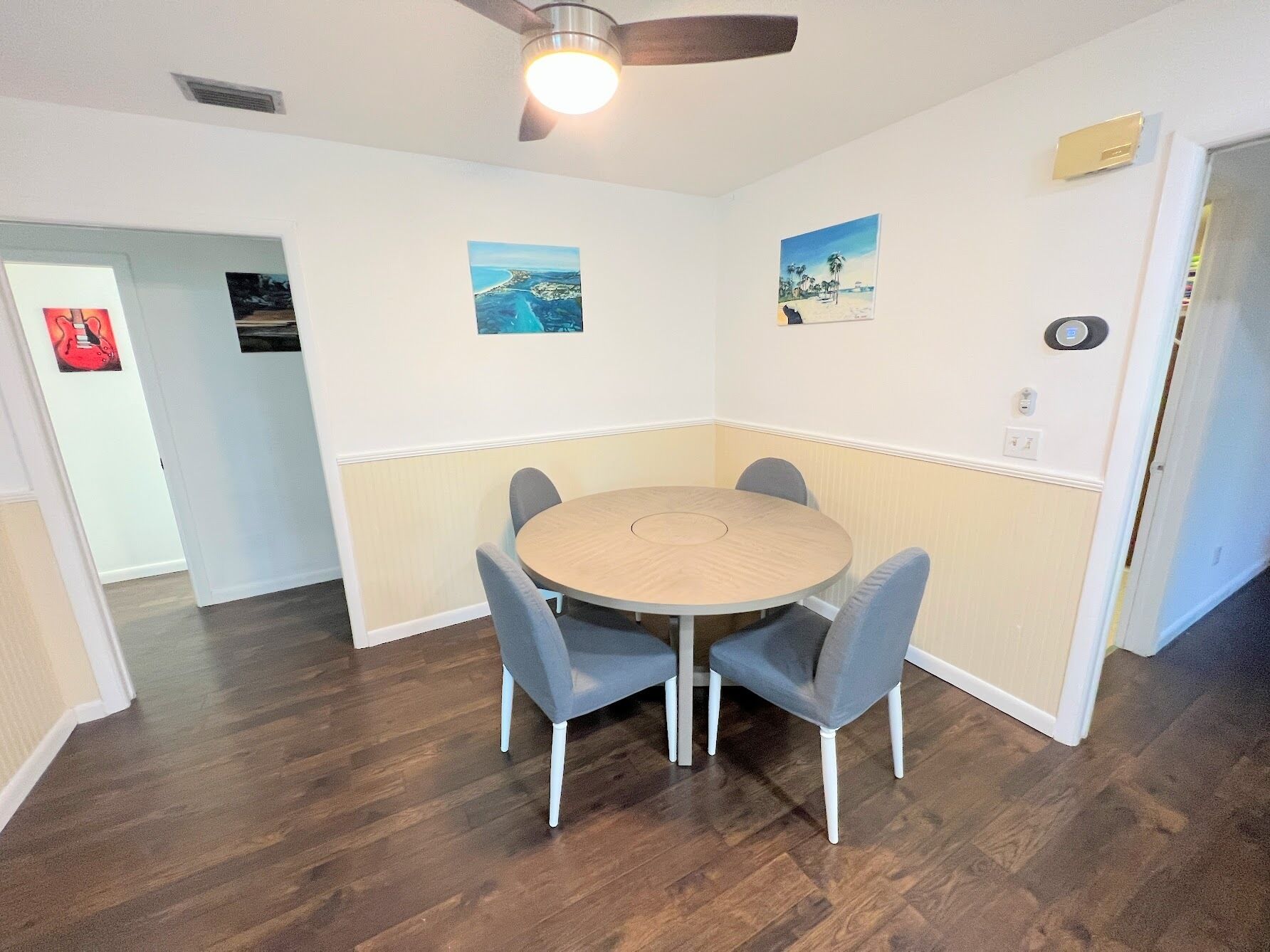A dining area with a round table and four chairs, a ceiling fan, and two wall-mounted paintings.