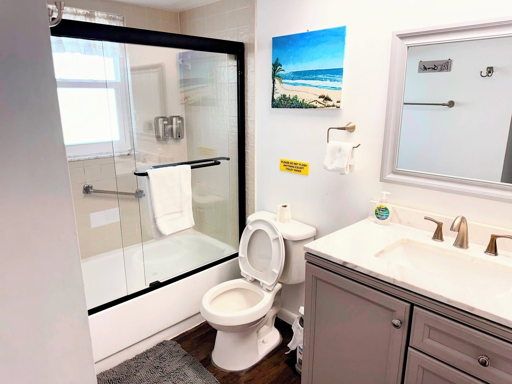 A modern bathroom with a glass shower enclosure, a toilet, a sink with a mirror, and a beach-themed painting on the wall.