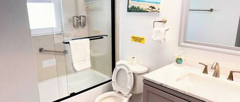 A modern bathroom with a glass shower enclosure, a toilet, a sink with a mirror, and a beach-themed painting on the wall.