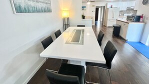 A modern dining area with a white table and black chairs, a painting on the wall, and a kitchen in the background.