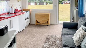Eine kompakte Küche mit weißen Schränken, einer roten Arbeitsplatte und einem kleinen Holztisch. Im Wohnbereich befindet sich eine graue Couch und ein Teppich mit Muster.