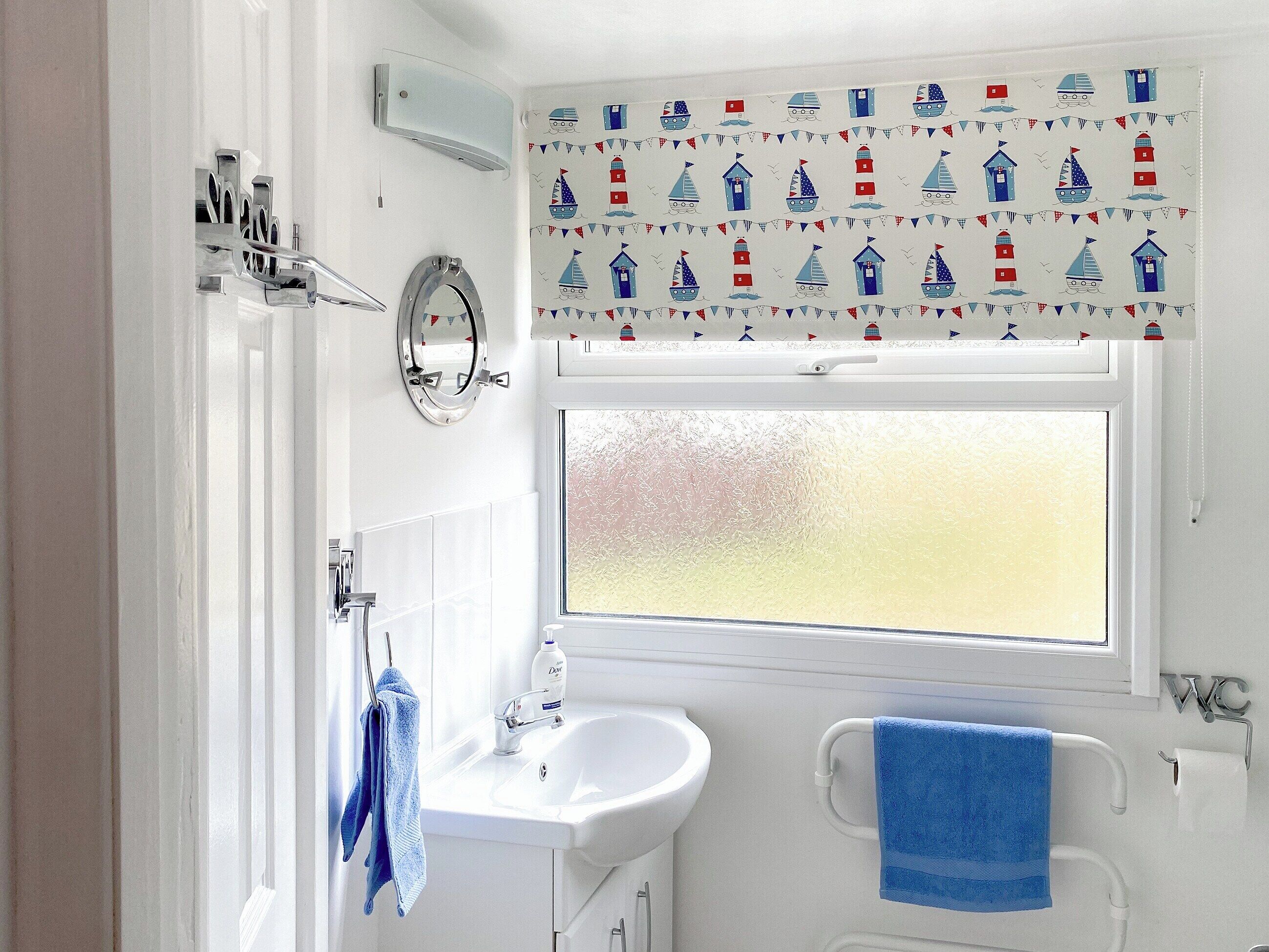 A bathroom with a white sink, mirror, and towel rack. There is a window with frosted glass and a curtain with nautical-themed designs.