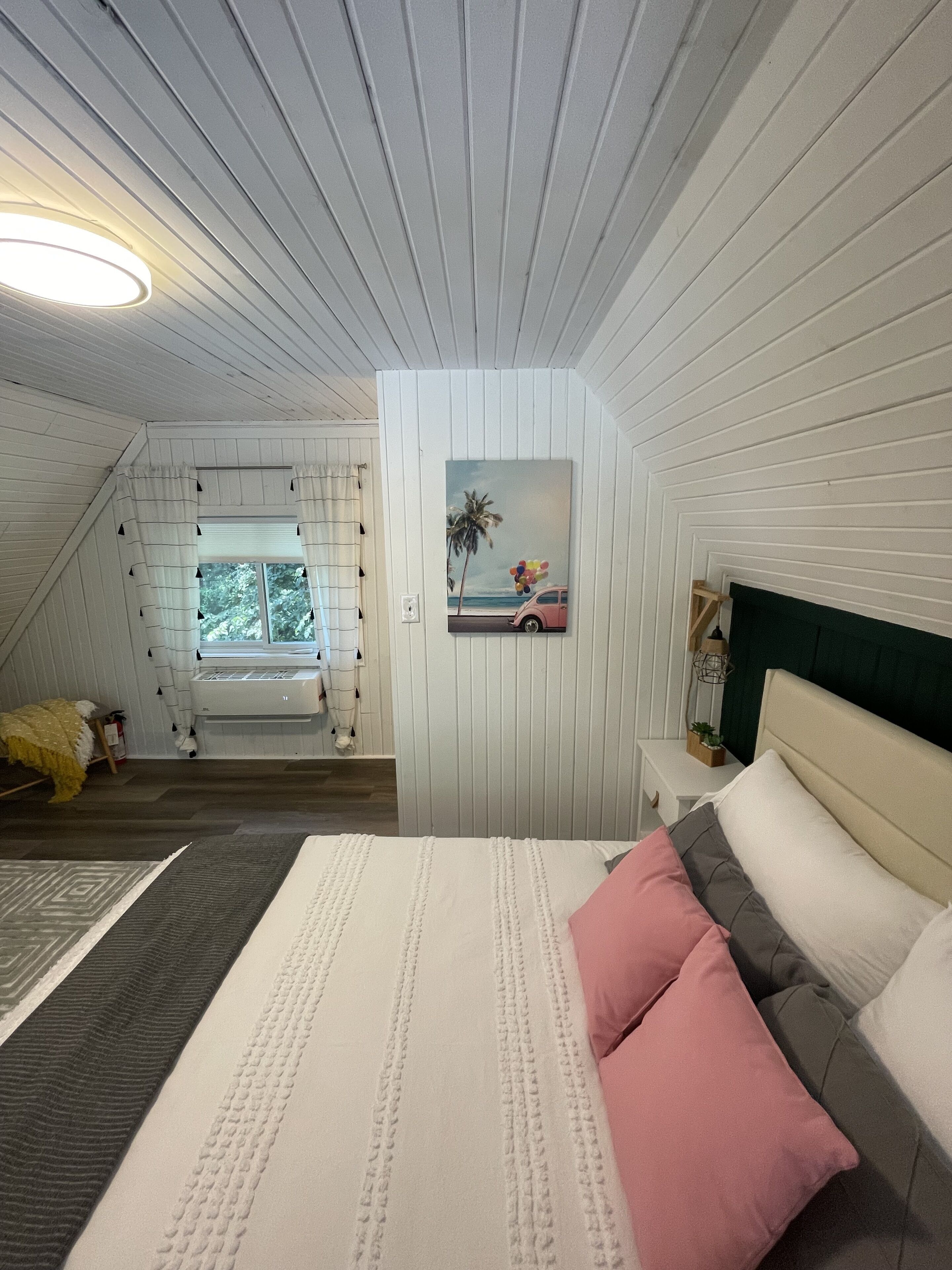 A bedroom with a bed, a nightstand, a window with curtains, a painting on the wall, and a ceiling light.