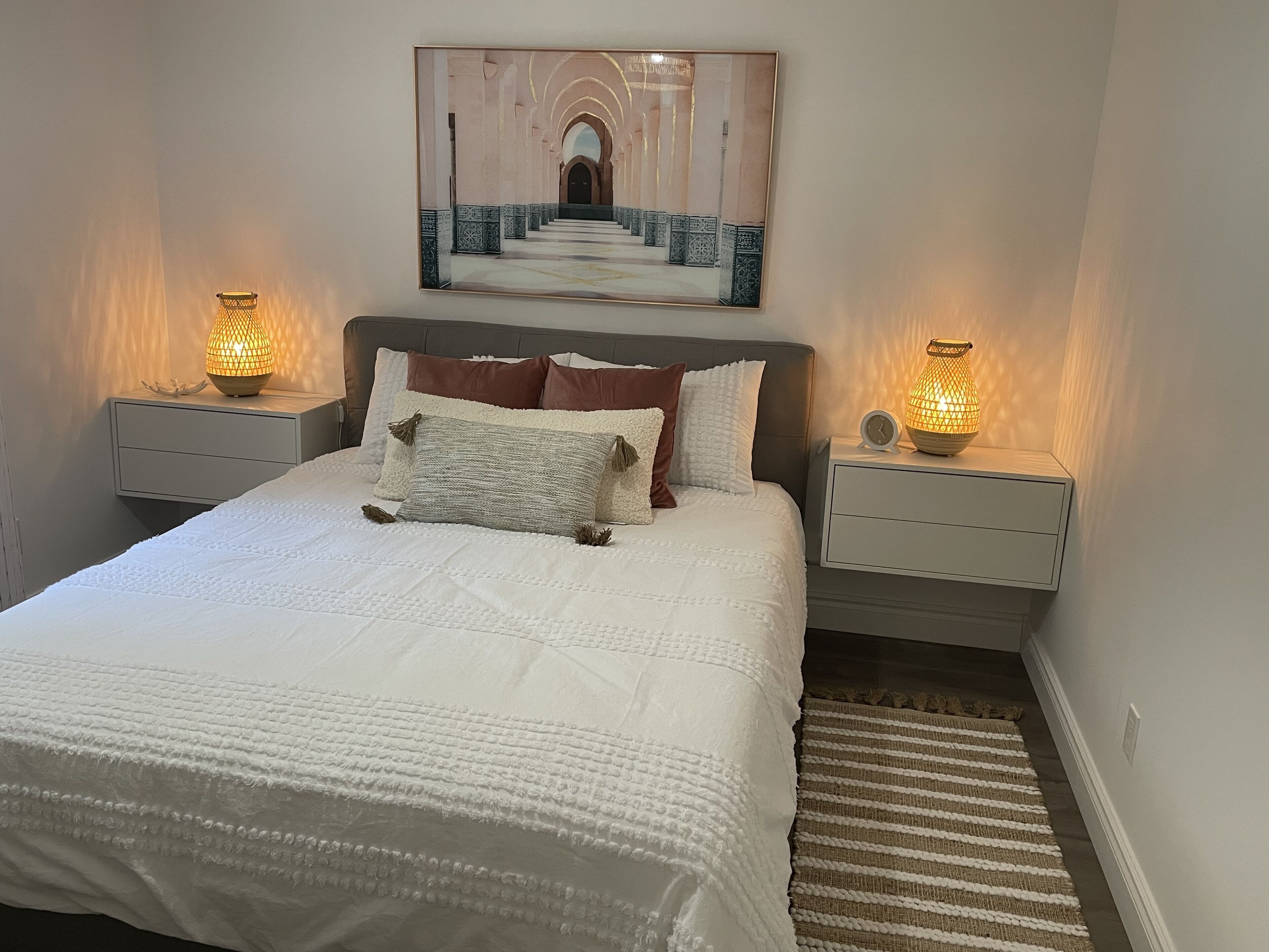 A neatly made bed with a white quilt, two nightstands with lamps, a framed artwork above the bed, and a striped rug beside the bed.