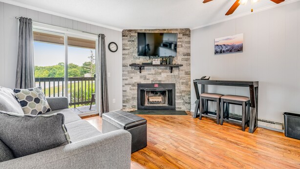 A living room with a fireplace, a TV mounted on the wall, a ceiling fan, and a balcony with curtains. - Gorgeous condo with hot tub, heated indoor pool, fireplace, balcony, & fast WiFi (Beech Mountain)