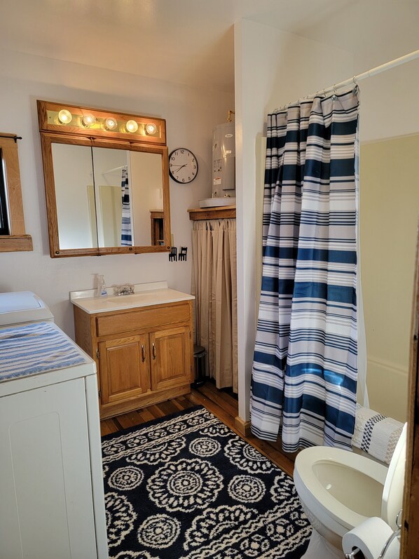 A bathroom with a wooden vanity, a mirror with lights, a clock, a striped shower curtain, and a patterned rug.