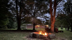 Een vuurkuil omringd door bomen in een grasrijke omgeving, met een huis op de achtergrond.