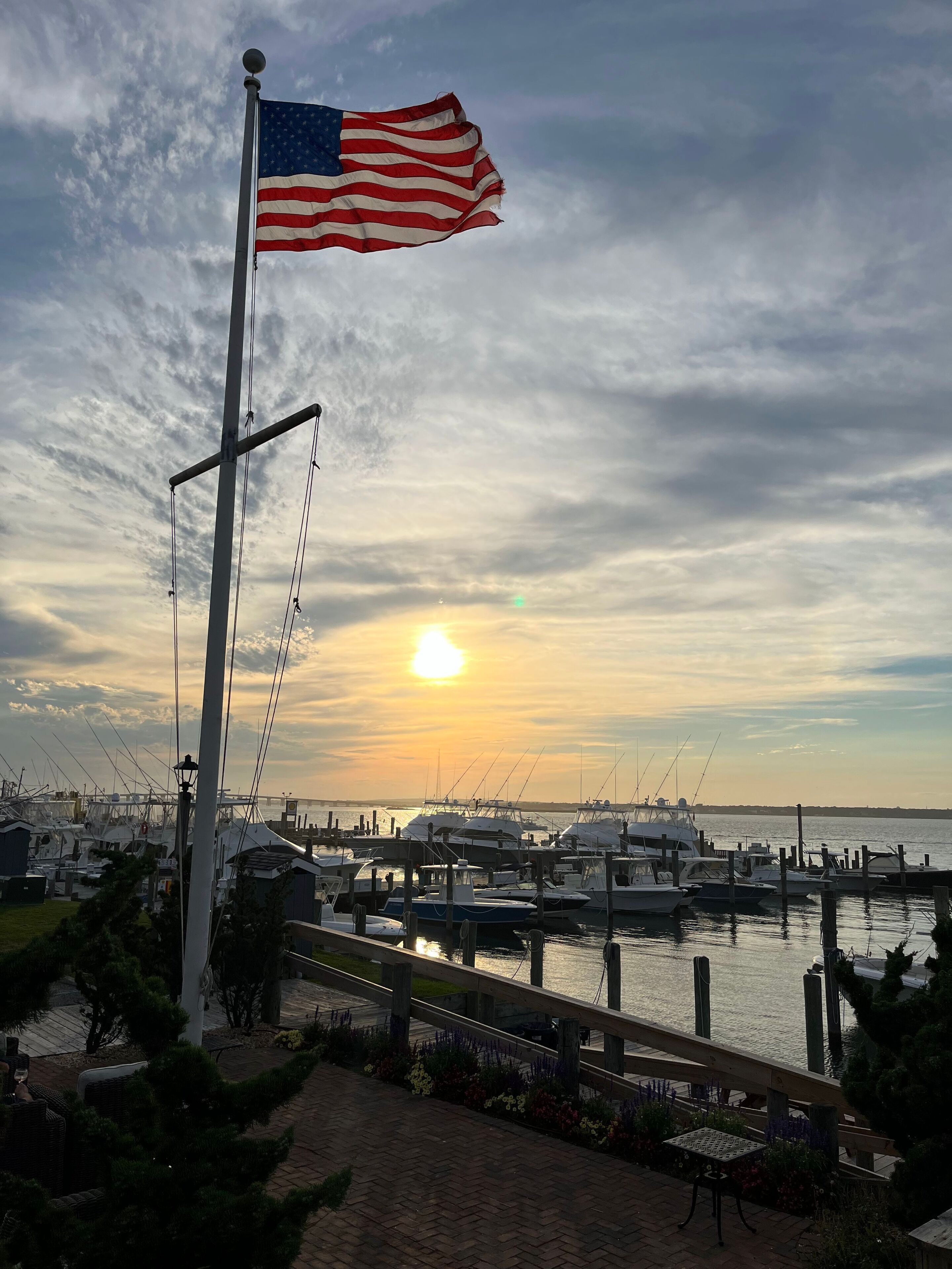 A flagpole with an American flag, a marina with boats, and a sunset in the background.