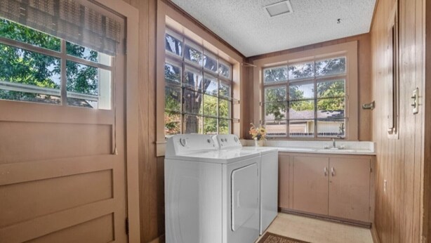 A laundry room with a front-loading washer and dryer, wooden cabinets, a sink, and large windows with a view of trees.