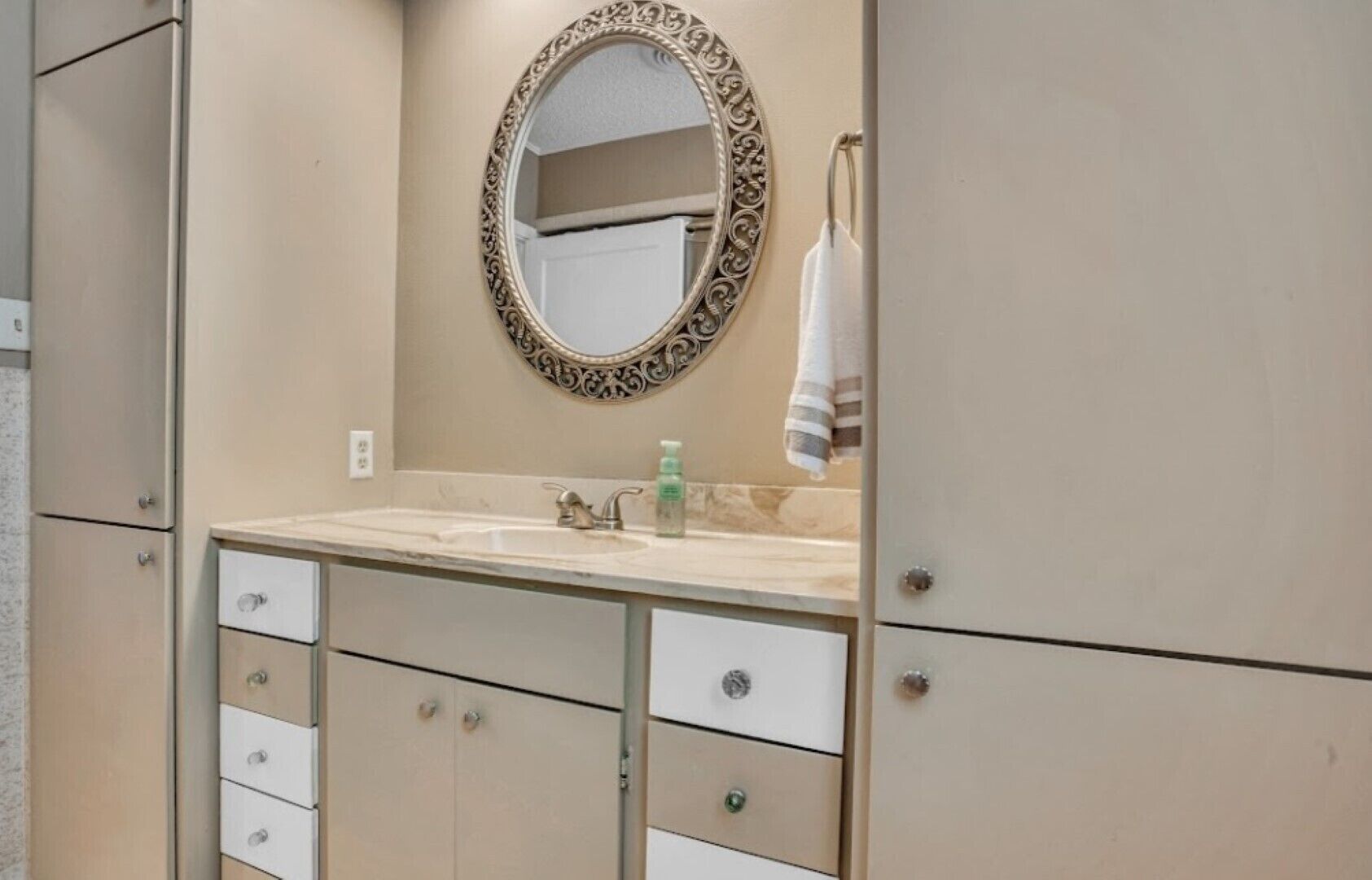 A bathroom with a beige vanity, a round mirror with an ornate frame, a sink with a faucet, and a towel hanging on a ring.