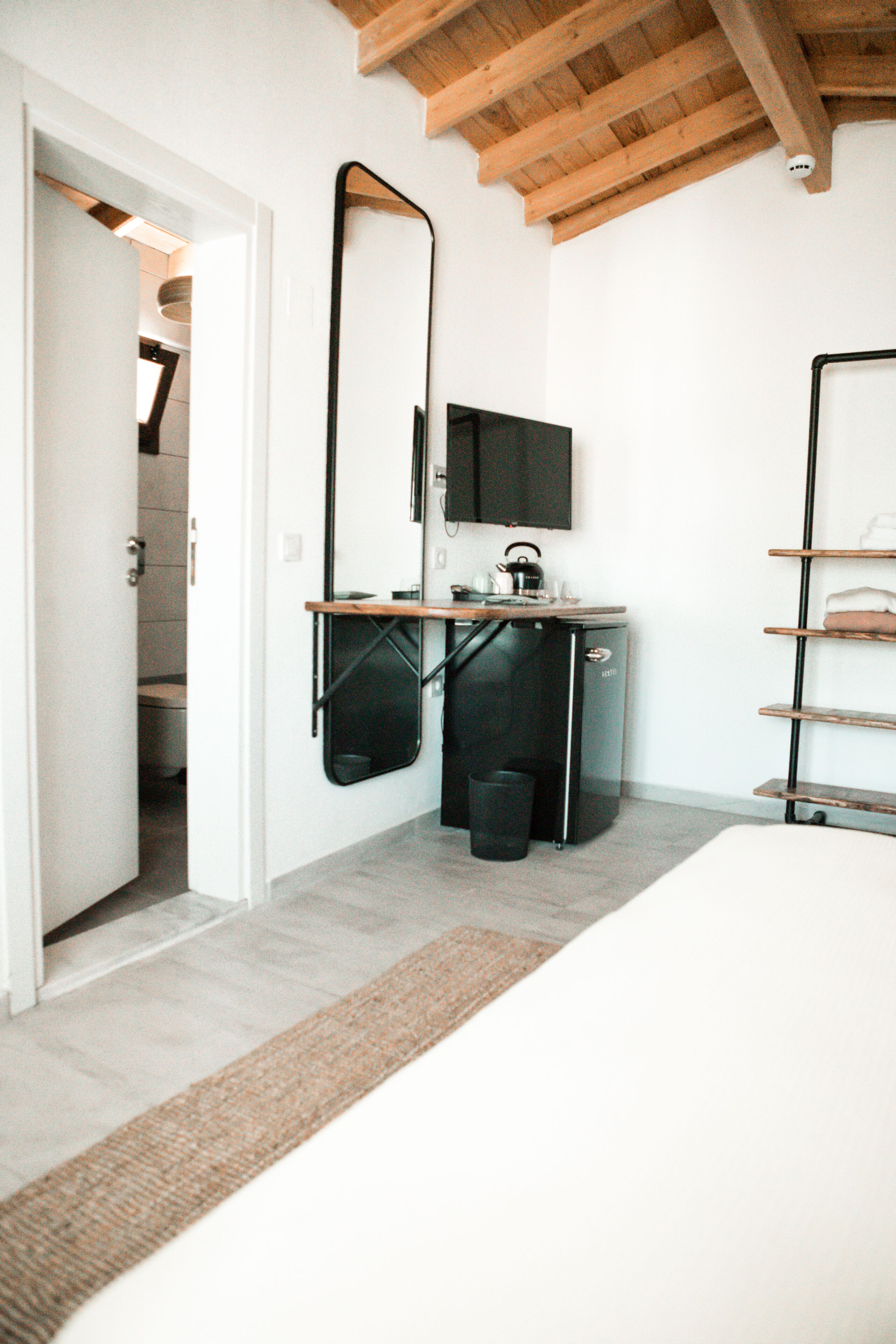 Un dormitorio compacto con una cama, un pequeño escritorio con hervidor, un espejo y una estantería.