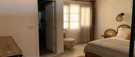 Un dormitorio con cama, techo de madera, ventana con cortinas y puerta que da a un baño.