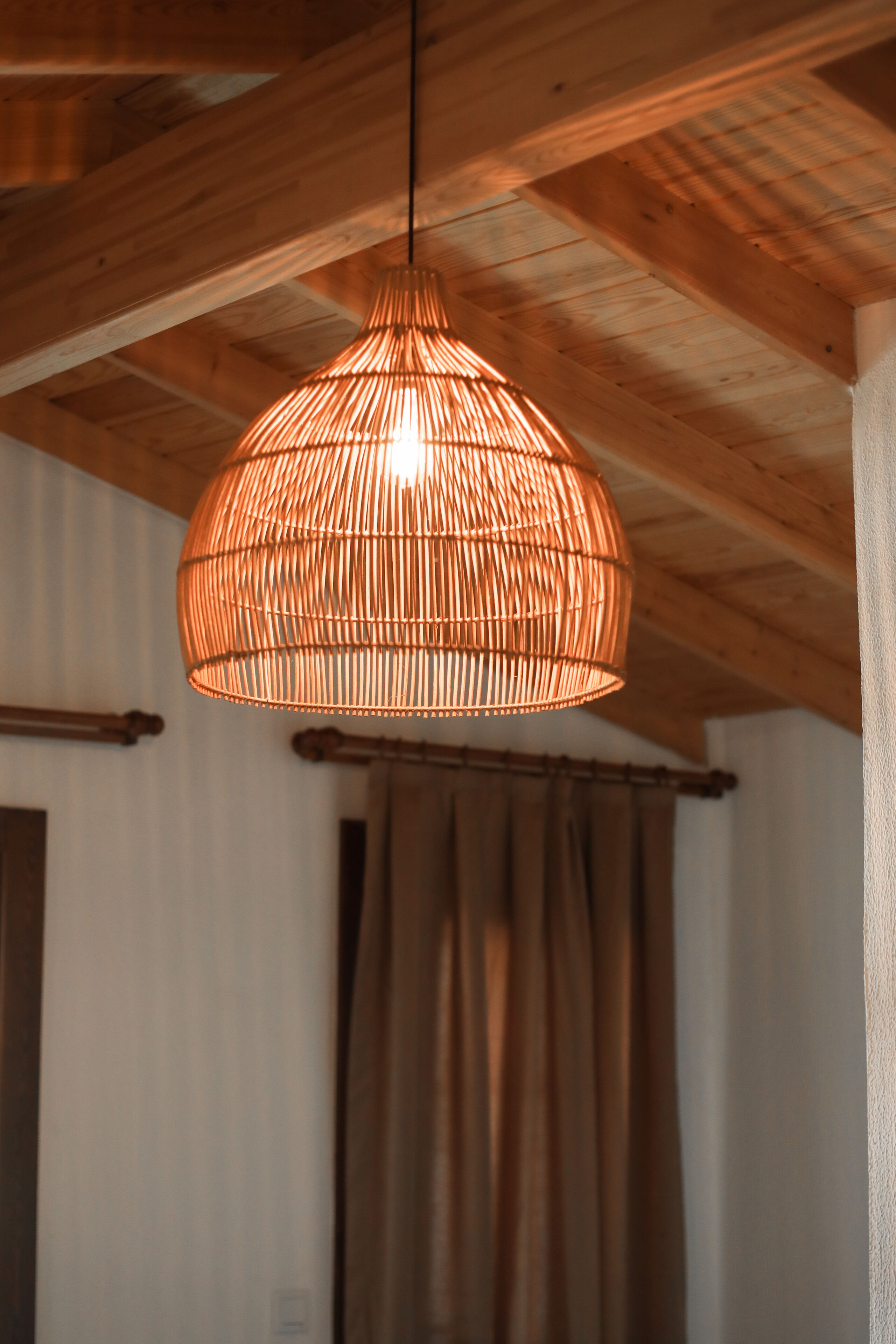 A room with exposed wooden beams, a woven rattan pendant light, and a curtain.