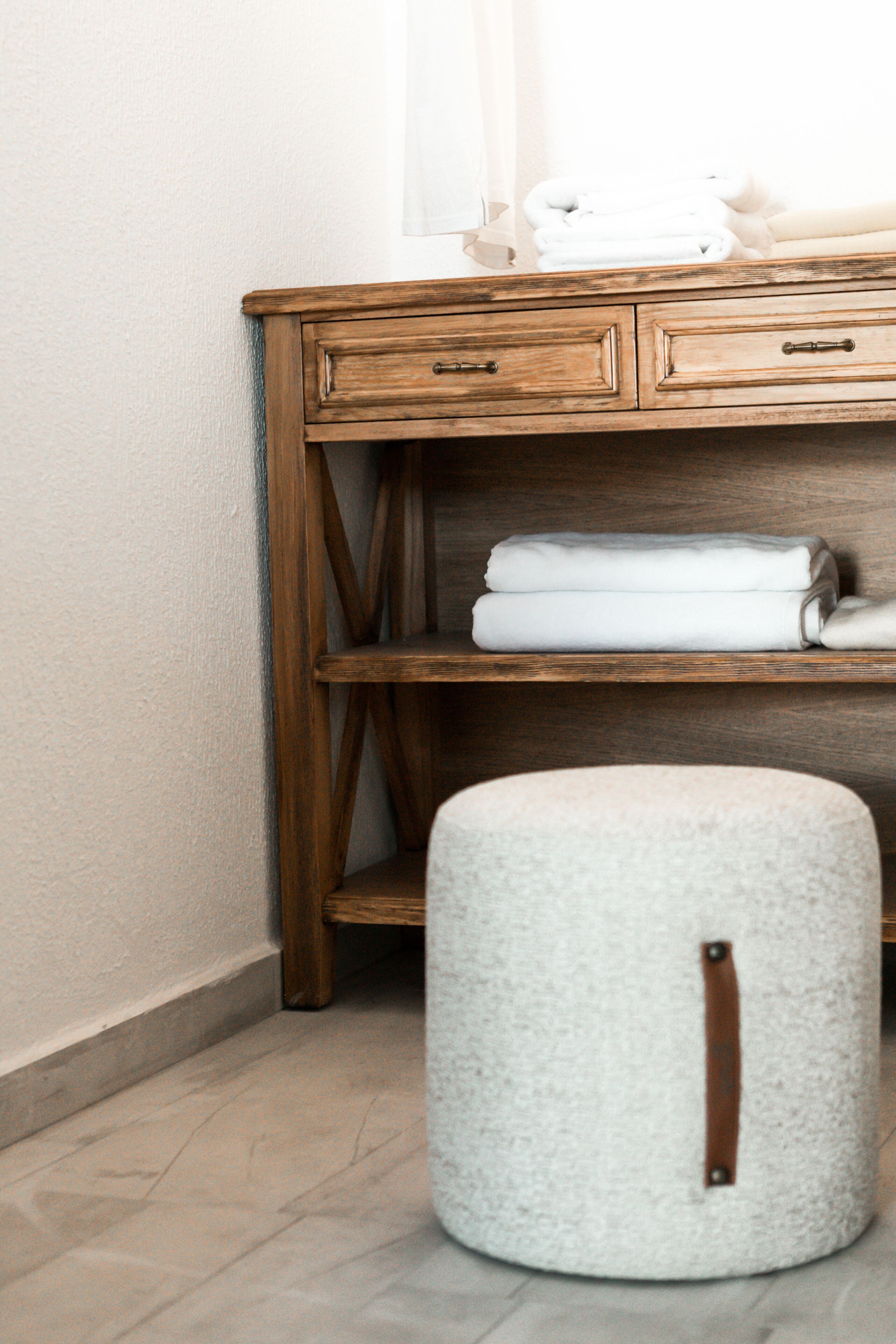 Une coiffeuse en bois avec une étagère où sont pliés des serviettes, et un pouf rond doté d’une sangle en cuir.