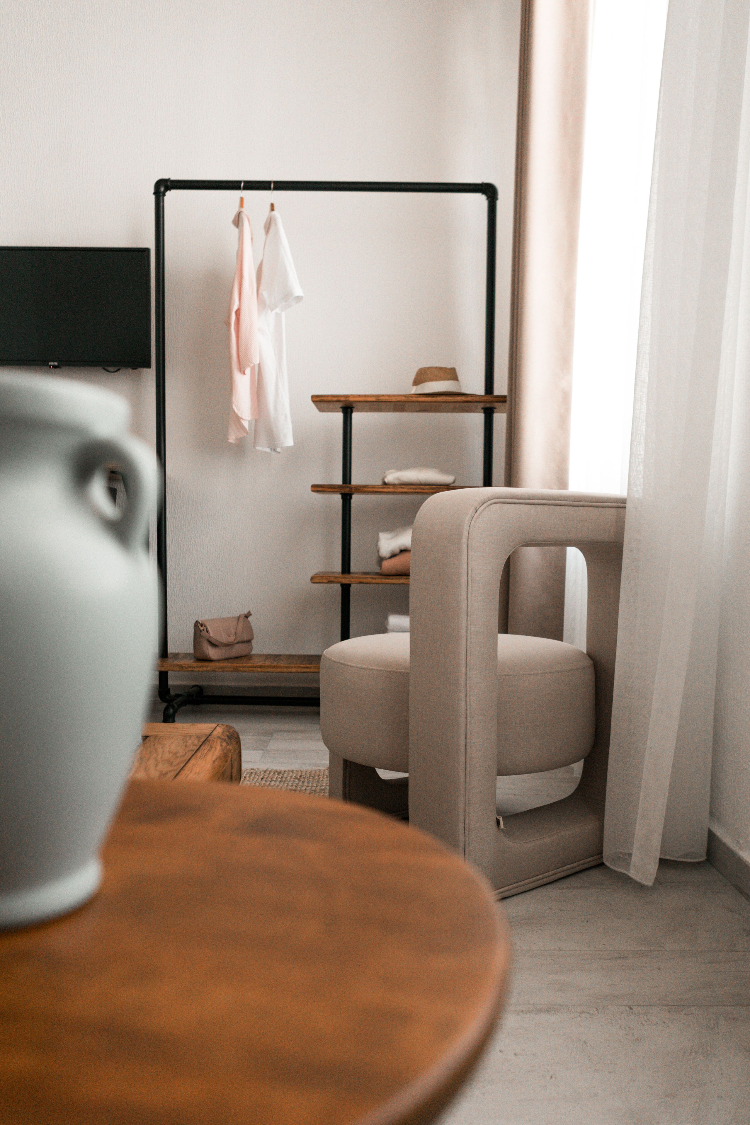 A modern hotel room with a minimalist design, featuring a wooden table, a comfortable armchair, a black TV mounted on the wall, and a clothing rack with hanging garments.