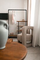 Habitación de hotel moderna con diseño minimalista, que incluye una mesa de madera, un sillón cómodo, un televisor negro montado en la pared y un perchero con ropa colgada.