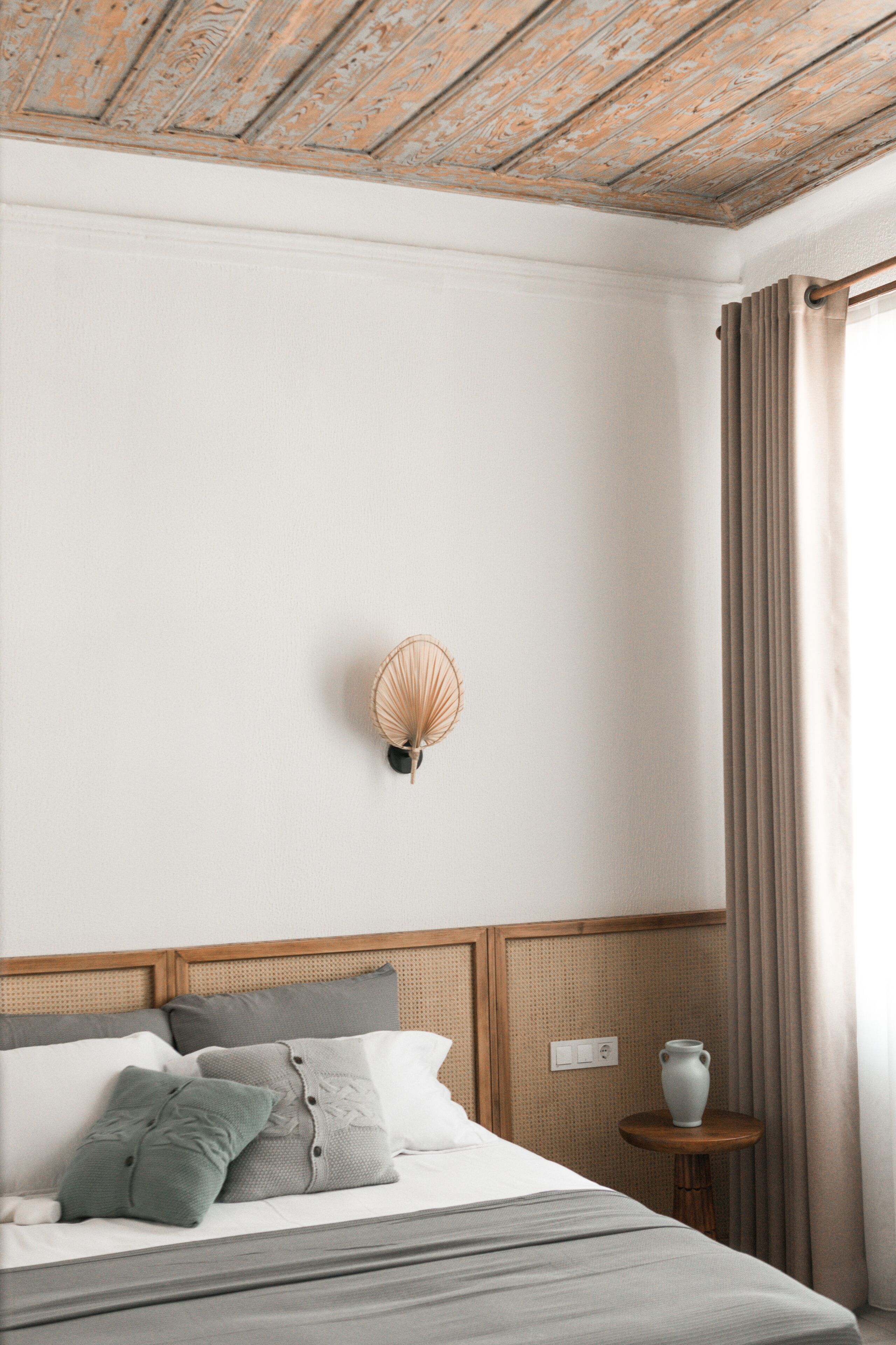 Una cama bien hecha con almohadas, una mesita de noche con un florero y una lámpara fijada a la pared.
