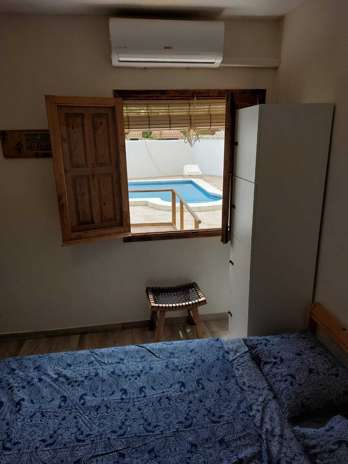 Quarto com cama, um banquinho de madeira, janela com persiana aberta e ar condicionado.