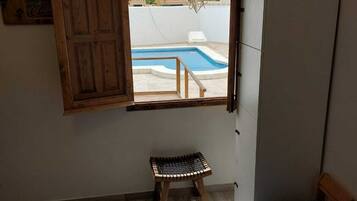 Un dormitorio con cama, un taburete de madera, una ventana con contraventanas abiertas y un equipo de aire acondicionado.