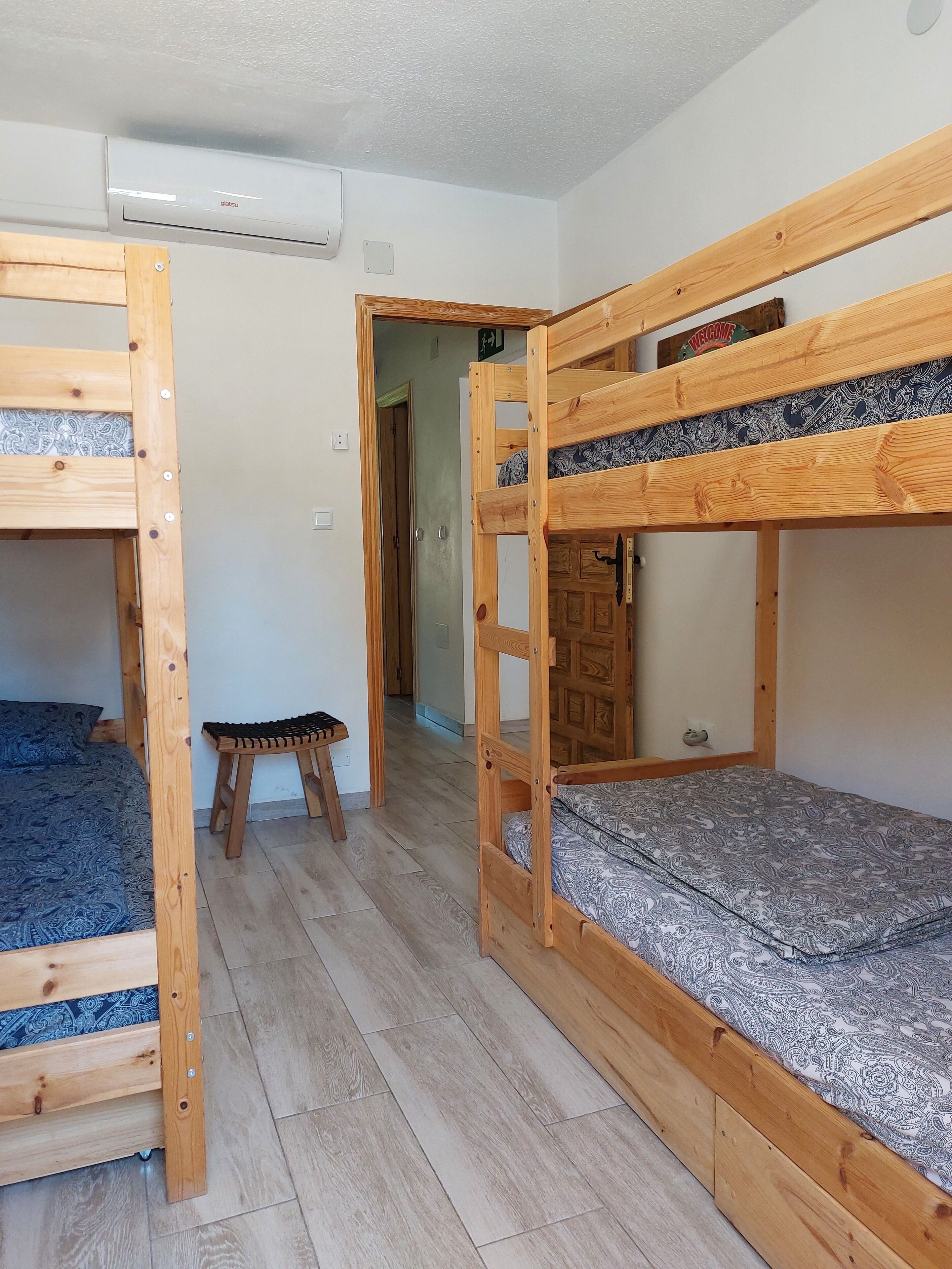 Una habitación con dos literas, un taburete de madera y una puerta que da a otra estancia.