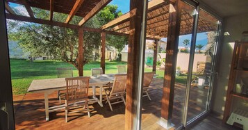 Un espacio habitable abierto con terraza de madera, puerta de cristal que da a un patio y vistas a un césped y árboles verdes.