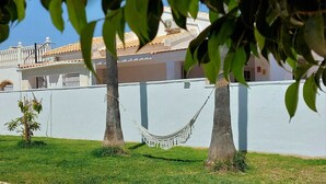 A hammock between two palm trees, a white wall, and a building with a tiled roof in the background. - ZENIA HOSTEL (Orihuela)