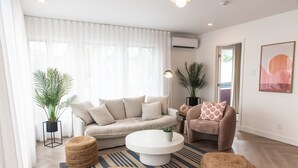 A modern living room with a beige sofa, a round coffee table, and two poufs. There are two potted plants, a floor lamp, and a framed artwork on the wall. - Hôtel Le Rosay (Venise-en-Quebec)