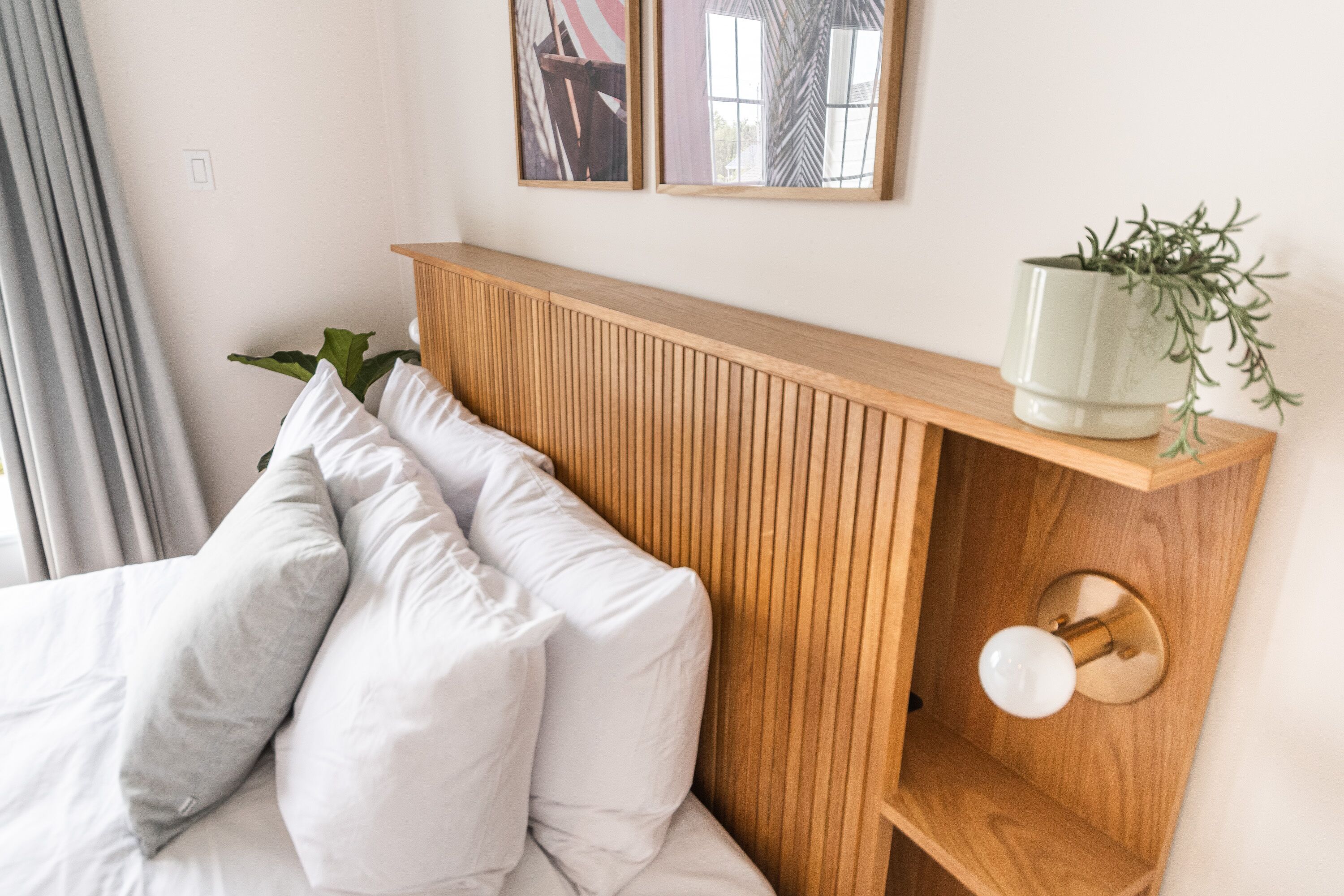 Una cama bien hecha con sábanas y almohadas blancas, cabecera de madera, planta de noche y lámpara de pared.