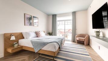 A modern bedroom with a wooden bed, a bedside table with a lamp, a large window with curtains, and a TV mounted on the wall.