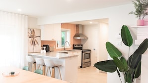 A modern kitchen with white cabinets, a central island with bar stools, stainless steel appliances, and a large green plant. - Hôtel Le Rosay (Venise-en-Quebec)