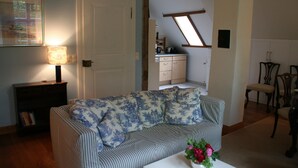 Un salon avec un canapé, une table basse ornée de fleurs, une lampe, une bibliothèque et, au fond, une cuisine.