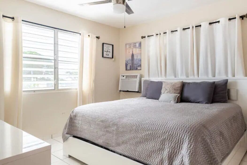 A bedroom with a bed, a window with curtains, a ceiling fan, and a wall-mounted air conditioner.