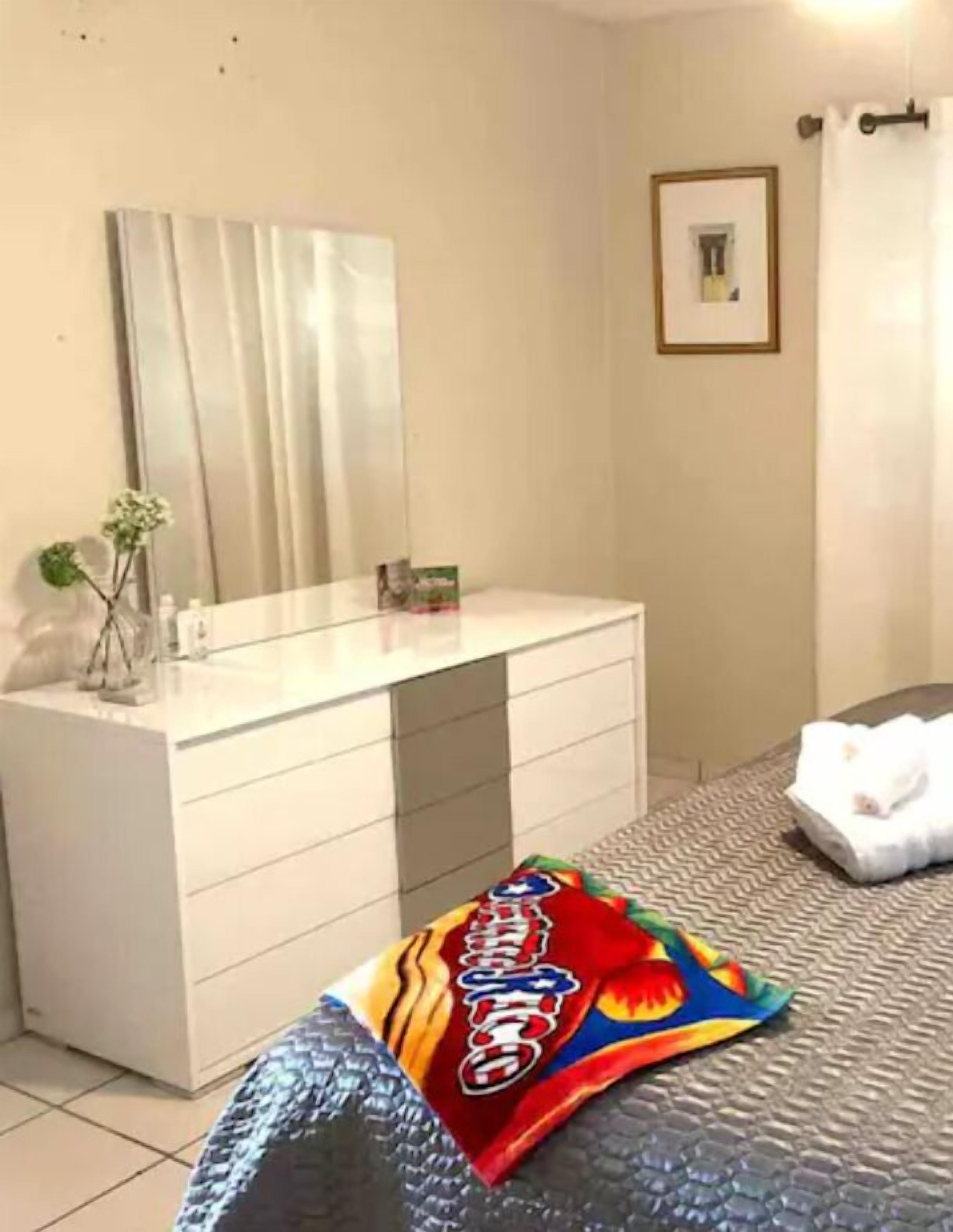A hotel room with a white vanity, a bed with a patterned bedspread, a framed picture on the wall, and a small plant on the vanity.