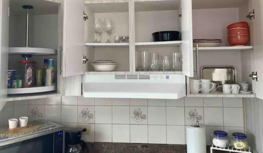Una cocina con armarios blancos, microondas y cafetera. Hay repisas con vasos y vajilla.