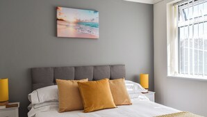 A neatly made bed with a grey headboard, two bedside lamps, and a painting of a beach scene above it.