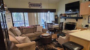 Uma sala de estar aconchegante com um sofá, cadeiras, uma mesa de centro e uma lareira. Há uma televisão montada acima da lareira e uma placa com a inscrição "Ski Chalet".