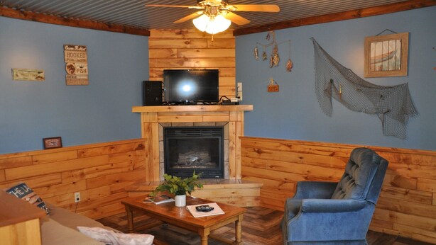 A cozy living room with a fireplace, wooden walls, a television, and a blue armchair. - 01) The Fishing Lodge (Bayfield)