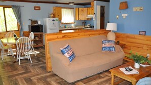 A cozy cabin-style room with a wooden floor, a sofa with pillows, a dining table with chairs, and a kitchenette with wooden cabinets. - 01) The Fishing Lodge (Bayfield)