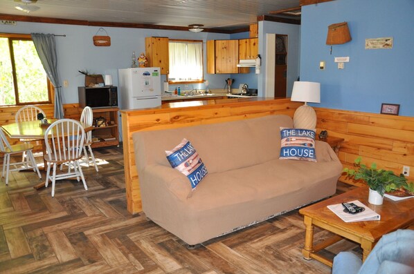 A cozy cabin-style room with a wooden floor, a sofa with pillows, a dining table with chairs, and a kitchenette with wooden cabinets.