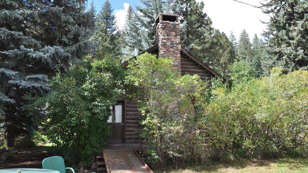 A rustic cabin with a stone chimney, surrounded by dense evergreen trees. - 04) The Rustic Lodge (Bayfield)