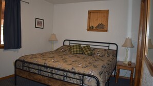 A single bed with a patterned bedspread, two bedside lamps, a framed picture on the wall, and a window with curtains. - 0005) The Wolf Cabin (Bayfield)